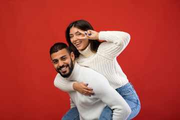Obraz premium A joyful Arab couple enjoys a playful moment together. The man gives his girlfriend a piggyback ride as she poses with a peace sign. They are clearly having fun against a vibrant red background.