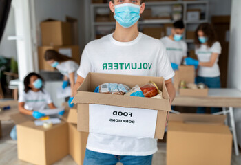 A male volunteer in a medical mask stands holding a box labeled food donations. Behind him, diverse colleagues prepare carton boxes in a charity organization setting, promoting community support.