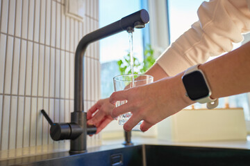 Hand filling glass with drinking water from kitchen faucet in modern home interior. Woman pouring...