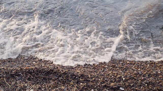 Waves crash onto pebbled shore, creating foam and spray. Dark pebbles, twigs, and seaweed scatter along the beach. Water recedes, leaving wet sand and debris behind. Natural coastal motion, quiet