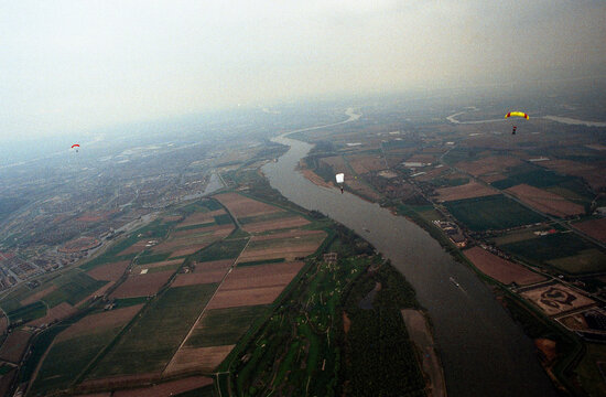 Scanned analog film aerial photograph of multiple skydivers with colorful parachutes descending over patchwork farmland and river with visible dust particles