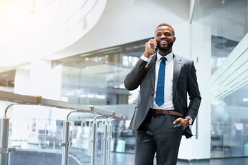 A man in a suit stands in a contemporary office space while talking on his phone. He smiles and...