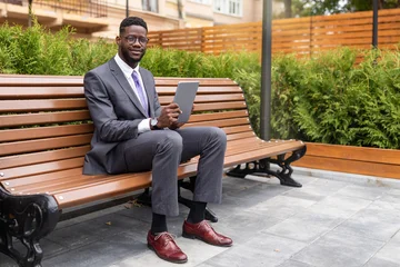 Fotobehang Ridders Happy black businessman sitting on bench in urban area and using digital tablet, websurfing, chatting or reading business news on pad, free space  © Prostock-studio