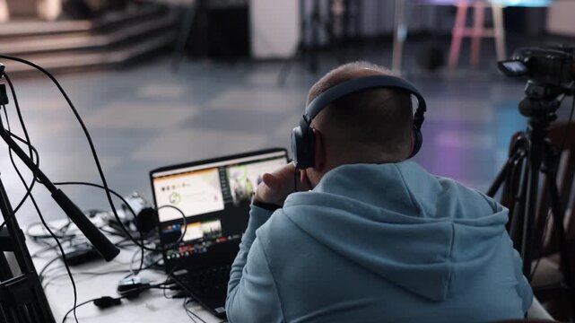 live streaming, video switcher, tripod camera. Man in headphones managing live video stream using laptop and camera setup during professional indoor event or conference.