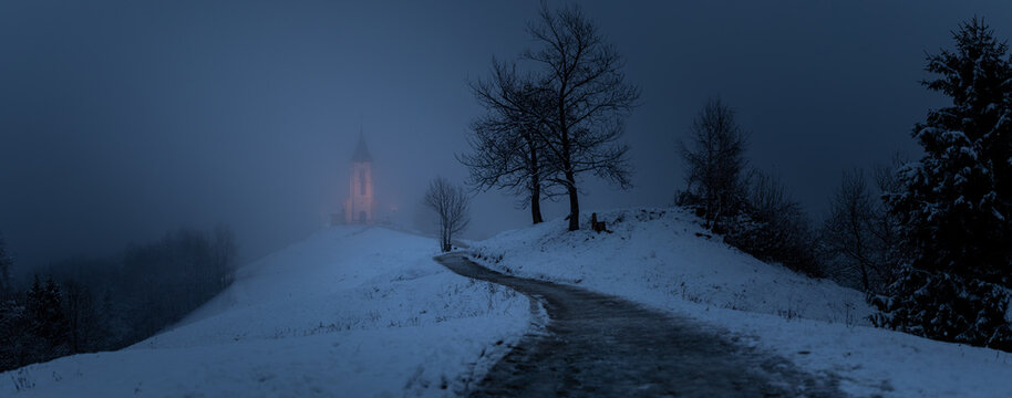 St. Primus und Felician bei D&auml;mmerung im dichten Nebel, beleuchtete Kirche auf verschneitem H&uuml;gel mit ansteigendem Weg und kahlen B&auml;umen, reduzierte Winterlandschaft mit k&uuml;hler Lichtstimmung