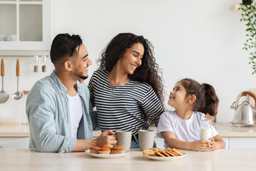 Cute happy arab family having breakfast while spending weekend together at home, cheerful young father, mother and curly little girl sitting at kitchen, drinking coffee and milk with homemade cookies