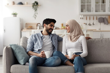 Fototapeta premium Fun together, enjoy breakfast and spare time at home, domestic date. Smiling attractive young arab lady in hijab and man talking and holding cups with empty space on sofa in living room interior