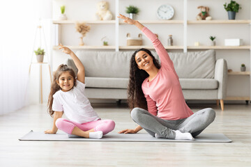 Sporty brunette mother and daughter sitting on fitness mat, stretching and smiling, home interior....