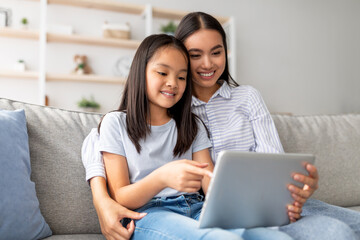 Fototapeta premium Excited asian mother and daughter using digital tablet together while sitting on sofa and surfing internet at home, free space. Happy mom and child playing online games on pad and hugging