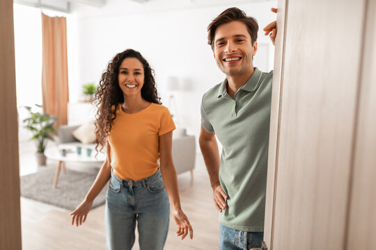 Cheerful young couple stands in the doorway of their modern flat, inviting guests inside with warm smiles. They are excited to host and eagerly await visitors to join them.