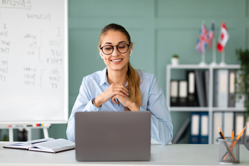 A cheerful female English teacher offers an online language class. She communicates on her laptop from her organized workspace, providing guidance and support to her students.