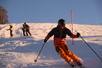 Action shot of a man skier skiing at fast speed on a snowy ski slope in the Alps. Orange suit, black skis, ski mask, and snow dust under sunset sky.