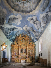 A Baroque altar with richly carved figures, a baptismal font in front; above, a painted wooden ceiling with a dramatic cloudscape, creating a sacred atmosphere
