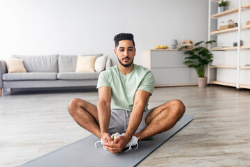 Home workout. Sporty young Arab guy stretching on mat in living room, practicing yoga, full length. Handsome Eastern man doing flexibility exercises, keeping fit and healthy during covid lockdown