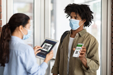 Young African American Man Showing Vaccination Certificate On Smartphone At Cafe Entrance, Female Cafeteria Worker In Medical Mask Using Digital Tablet For Screening Health Qr Code Of Customer