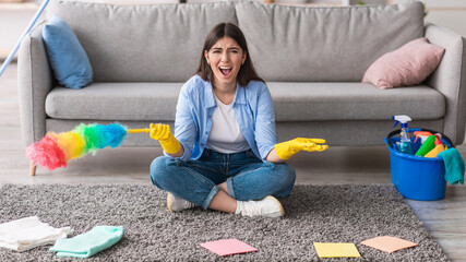 Tired lady screaming while cleaning her apartment, wearing rubber gloves and holding feather...