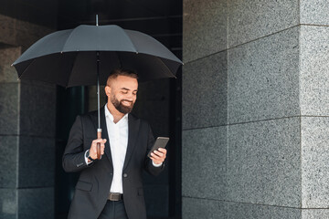 Happy handsome mature businessman using smartphone while holding black umbrella during walk in the city on rainy day. Man texting or surfing internet outdoors
