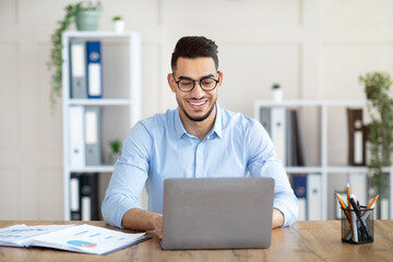 Fototapeta premium Portrait of confident young Arab guy sitting at his table with laptop computer, typing on keyboard in office. Millennial businessman using portable pc for online job, having virtual meeting