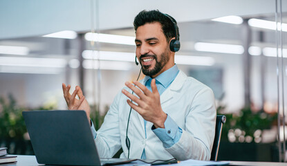 A doctor is having an online consultation with a patient. He is smiling and wearing a headset while sitting at a desk with a laptop in a modern office. Plants can be seen in the background.
