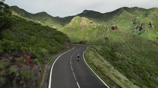 A lone cyclist navigates a winding mountain road through a vibrant green, hilly landscape under an overcast sky, embodying outdoor adventure, fitness, and exploration of nature's beauty.
