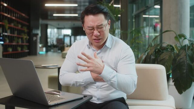 Stressed Chinese mature businessman using laptop and suffers from tension holding chest with heart pain attack risk of stroke symptom while working in modern office lobby. Businessperson in shirt.