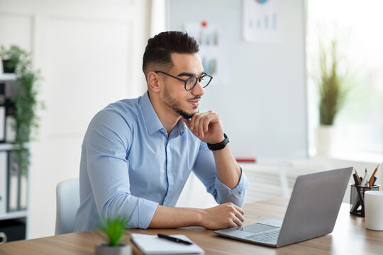 Positive Arab businessman using laptop pc at workplace, speaking to colleagues on webcam. Successful millennial office worker reading important document online, checking email