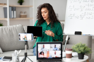E-Learning. Confident woman having video conference call using laptop. Female teacher showing digital tablet, recording tutorial with mobile phone on tripod, streaming live to diverse students