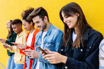Obraz premium Happy group of young multiracial friends using smart phones together outside over yellow background. Social media and technology lifestyle concept