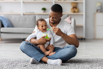 Happy Young Black Dad Spoon Feeding His Cute Adorable Baby Son At Home, Loving African American Father Taking Care About Little Male Infant Child Enjoying Paternity Leave Time, Free Space