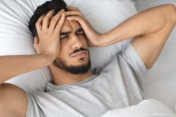 A young Arab man is lying in bed, struggling with an acute headache. He touches his temples,...