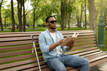 Obraz premium Young black visually impaired man sitting on bench in city park, reading Braille book outdoors. Millennial blind guy in dark glasses studying or enjoying entertainment literature, outside