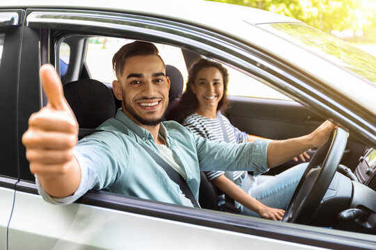 Handsome arab guy sitting inside car with gilfriend and showing thumb up, smiling at camera. Happy middle-eastern couple or family renting auto for their weekend trip, going vacation together