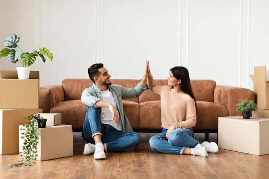 Good Job Concept. Happy young couple tenants giving high five, celebrating moving day, sitting on the floor near carboard boxes and plants. Smiling man and woman enjoying relocation