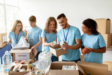 Group of young volunteers from diverse backgrounds pack canned food and other products into cardboard boxes for a charitable foundation. They work together in a bright, organized space.
