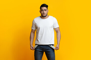 Portrait of sad middle-eastern young man showing empty pockets on yellow colored studio background,...