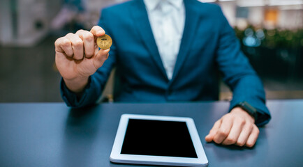 A person in a blue suit holds a crypto coin in one hand. A tablet is on the table in front of him. The setting appears to be a modern office space with indoor lighting.
