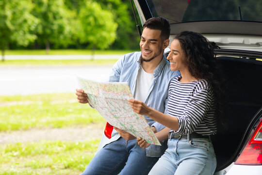Loving middle-eastern couple watching road map while having break, travelling by car together, panorama with copy space. Smiling arab man and pretty ady sitting on open auto trunk, checking location