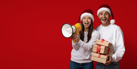 A joyful couple dressed in festive outfits holds a megaphone and presents. They are celebrating the Christmas season with smiles on their faces and red backgrounds.