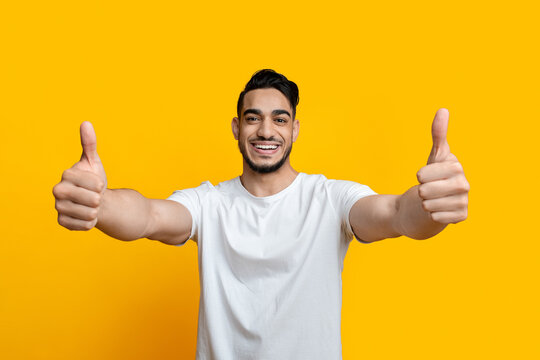 Happy young arab man showing thumbs up at camera over yellow studio background, cheerful handsome middle-eastern guy recommending or definitely like something, panorama with copy space