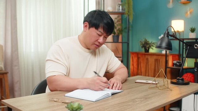 Asian man sitting at home table writes budget notes, crossing numbers again, rewriting lines. Chinese guy frowns, tapping pen, showing stress while planning financial decision and seeking solution.