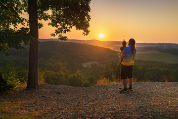 Rear view of a father holding his toddler, sharing a peaceful bonding moment during a golden sunset over the hills.