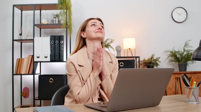 Middle-aged businesswoman at home office folds hands in prayer at laptop looks up hopes for approval. Freelancer girl at table waits for reply checks inbox breathes writes note stays calm faith inside