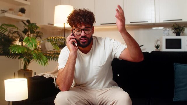 Arabian man at home answers smartphone gets spam call from pushy operator says no feels angry annoyed. Egyptian guy on sofa rejects fake offer blocks number sighs frowns frustrated after fraud call.