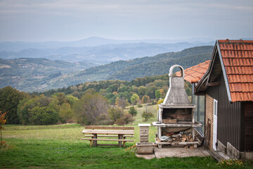 Outdoor barbecue and wooden bench set in a rural Serbian landscape with hills, trees and open nature, showing a peaceful countryside cooking and leisure area.