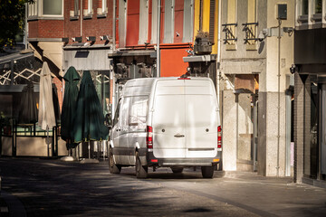 A white delivery van driving down a paved urban street lined with shops in the city of Heerlen, The Netherlands, a symbol of business and logistics, last mile delivery.