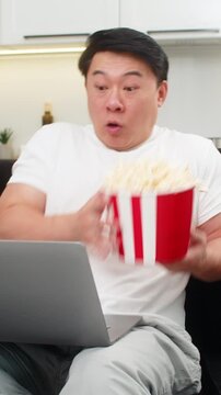 Asian man at home watches scary thriller on laptop jumps at scare spills popcorn then breathes and slows pulse. Chinese guy on sofa keeps watching curious holds backet firm and waits for big twist