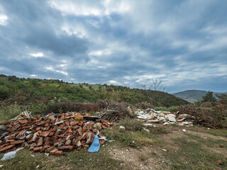 Illegal garbage dump with construction debris in a rural landscape in Serbia. Piles of bricks and waste litter the grassy field contrasting with the green hills and serbian nature.