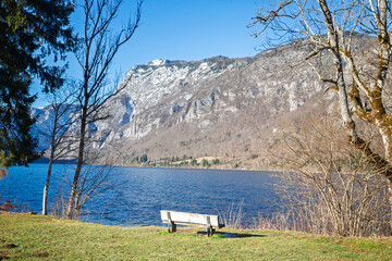 Empty bench on the shore of Lake Bohinj in Slovenia facing the Julian Alps, with deep blue water, bare trees and snow on high ridges, creating a peaceful winter panorama and travel scene.