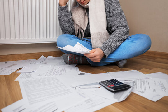 A man in a scarf sits on the floor with utility bills and a calculator next to the radiator, worried about high heating costs.