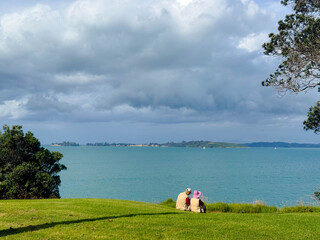An asian old couple is sitting on green grass field and looking at beautiful blue ocean in New Zealand.